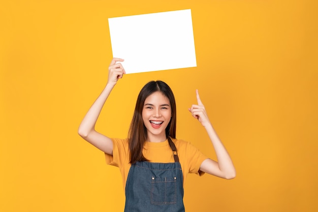 Happy young Asian woman wears an apron and holding a blank paper with smiling face and looking at the orange background. for advertising signs.