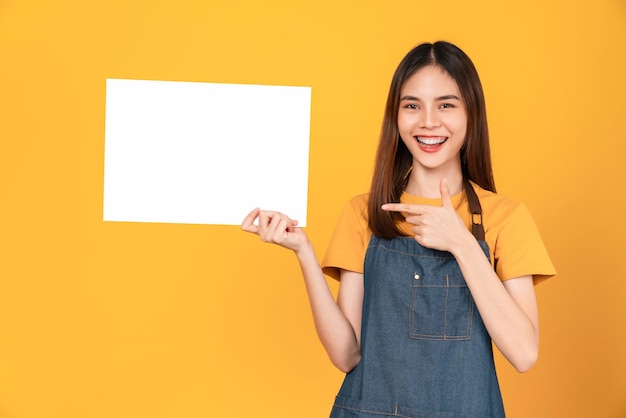 Happy young Asian woman wears an apron and holding a blank paper with smiling face and looking at the orange background. for advertising signs.