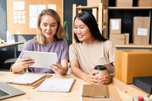 Happy young Asian woman with coffee sitting by table next to colleague with tablet while both looking through new online orders of clients