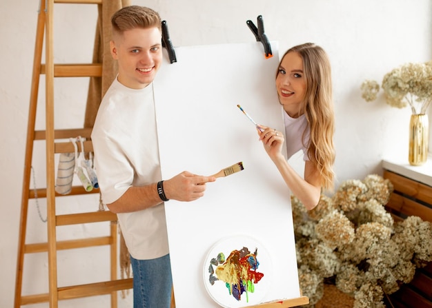 Happy young attractive couple stand behind easel with blank canvas and watercolor palette posing hol...