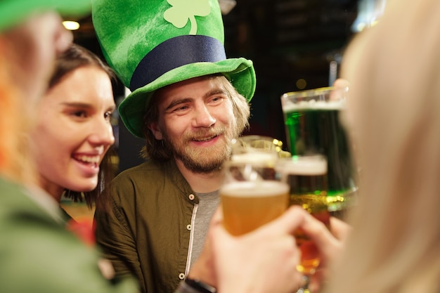 Happy young bearded man in traditional irish hat holding glass of beer and looking at one of his friends while toasting for saint patrick day