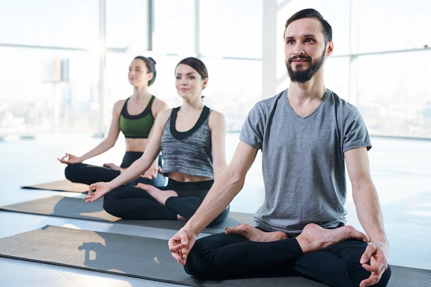 Happy young bearded man and two girls in sportswear sitting in pose of lotus on mats while repeating after instructor