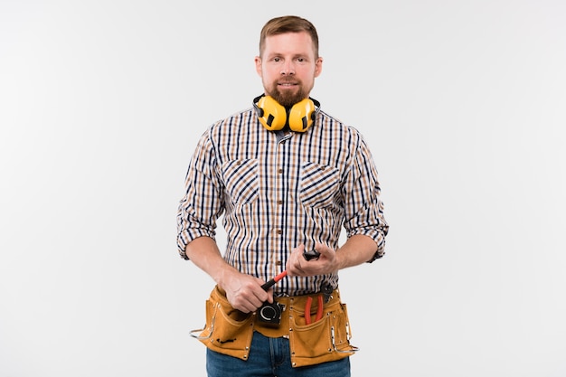 Happy young bearded repairman with protective earphones, hammer and tool belt standing in front of camera in isolation