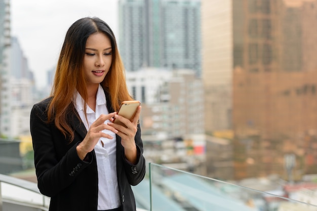 Happy young beautiful Asian businesswoman using phone against view of the city