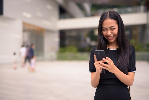 Happy young beautiful Asian tourist woman using phone at the mall outdoors