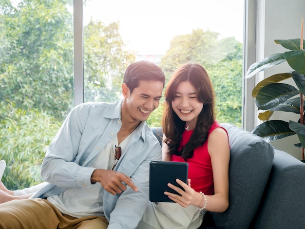 Happy young beautiful Asian woman and handsome man smile together while looking at tablet screen on grey sofa near the glass window at white living room Couple lover using digital tablet together