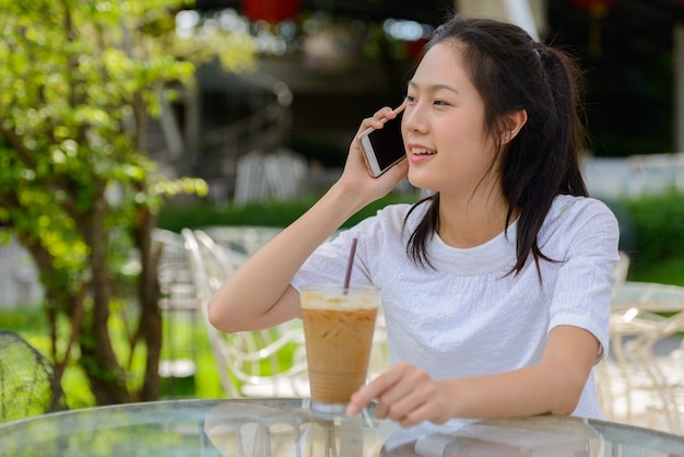 Happy young beautiful Asian woman talking on the phone at the coffee shop outdoors