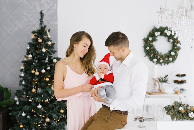 A happy young beautiful family with a young son in a Santa costume hold him in their arms in the living room decorated for Christmas