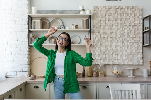 Happy young beautiful woman in glasses and green shirt dancing and singing at home in the kitchen