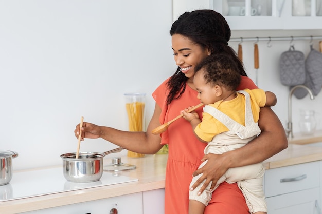 Happy Young Black Mother Cooking In Kitchen With Little Baby On Hands