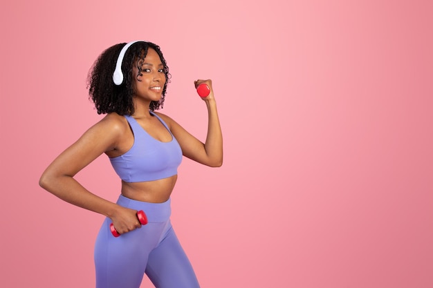 Happy young black woman in sportswear and wireless headphones doing hand exercise with dumbbells