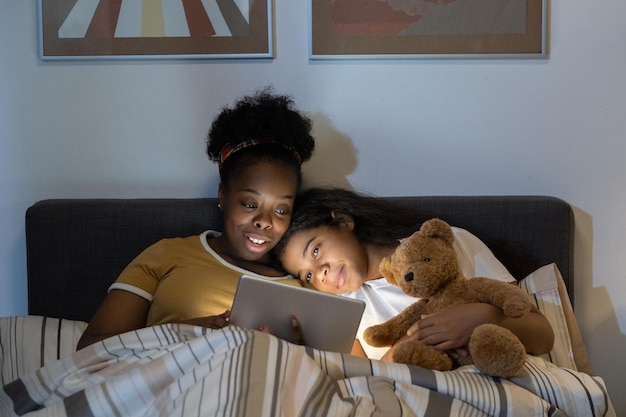 Happy young blackwoman with afro hairstyle lying under duver in bed and using tablet