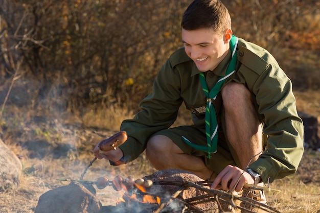 Happy Young Boy Scout Grilling Sausages For Food to Eat on the Ground at the Camp Area.