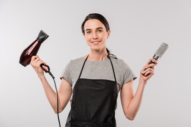 Happy young brunette hairdresser in apron holding hairdryer and hairbrush while standing in front of camera in isolation