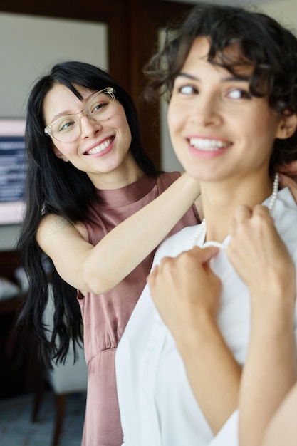 Happy young brunette woman helping her sister put pearl necklace on neck