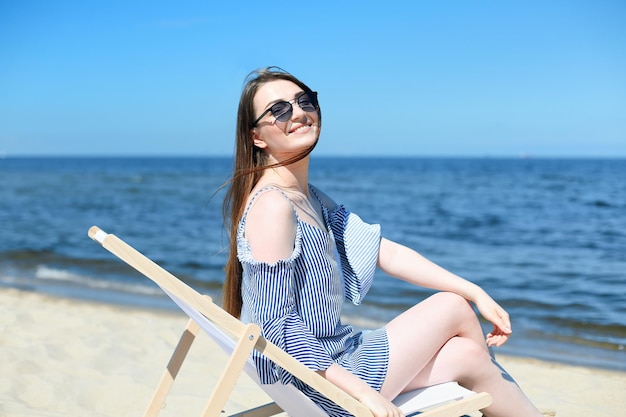 Happy young brunette woman relaxing on a wooden deck chair at the ocean beach while looking at camera, smiling, and wearing fashion sunglasses. The enjoying vacation concept