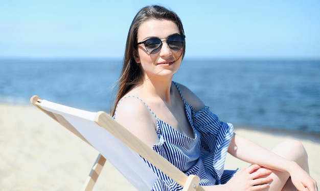 Happy young brunette woman relaxing on a wooden deck chair at the ocean beach while looking at camera, smiling, and wearing fashion sunglasses. The enjoying vacation concept