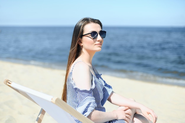 Happy young brunette woman relaxing on a wooden deck chair at the ocean beach while smiling, and wearing fashion sunglasses. The enjoying vacation concept