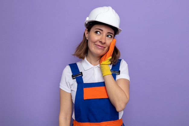 Happy young builder woman in construction uniform and safety helmet in rubber gloves looking aside smiling touching her cheek