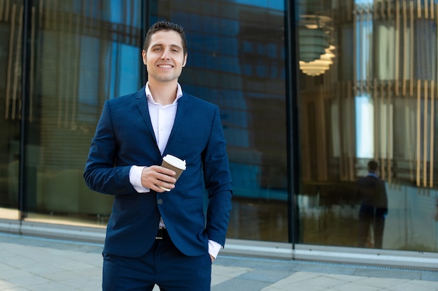 Happy young business man smiling and holding cup of coffee in front of modern building