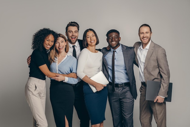 Happy young business team in formalwear embracing while standing on beige background