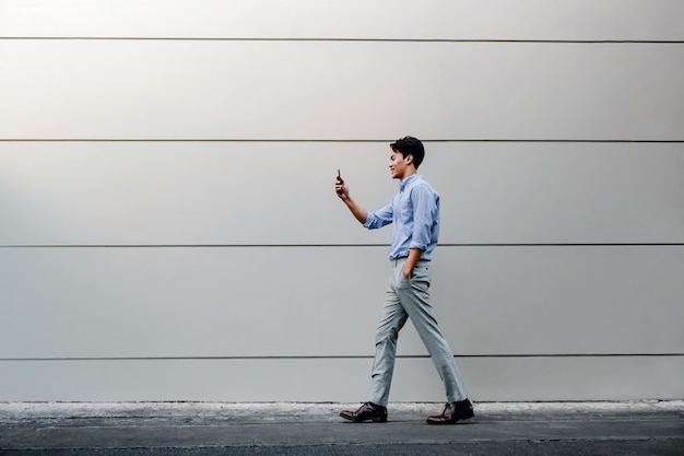 Happy Young Businessman in Casual wear Using Mobile Phone while Walking by the Urban Building Wall. Lifestyle of Modern People.