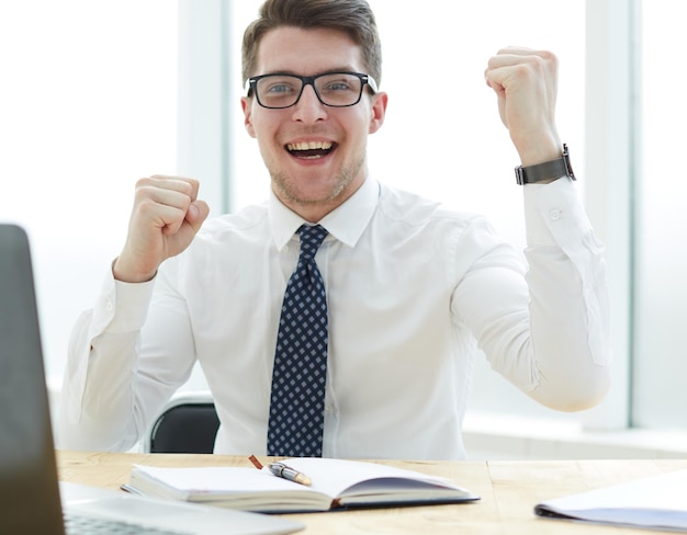 Happy young businessman raising hands in front of laptop at office desk