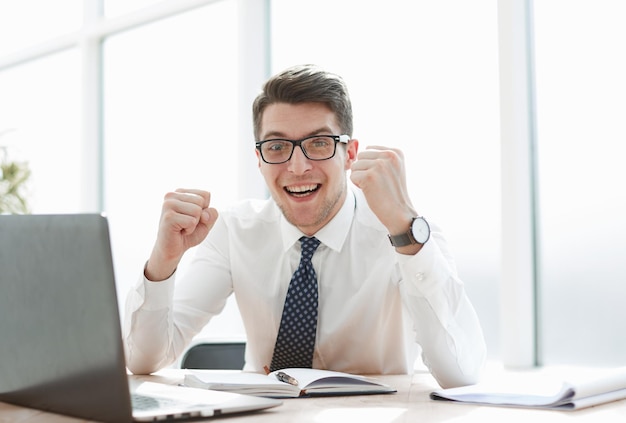 Happy young businessman raising hands in front of laptop at office desk