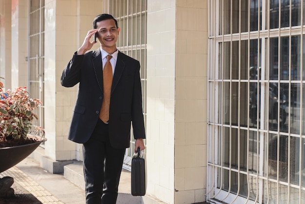 Happy young businessman with briefcase walking and talking on cell phone near business center