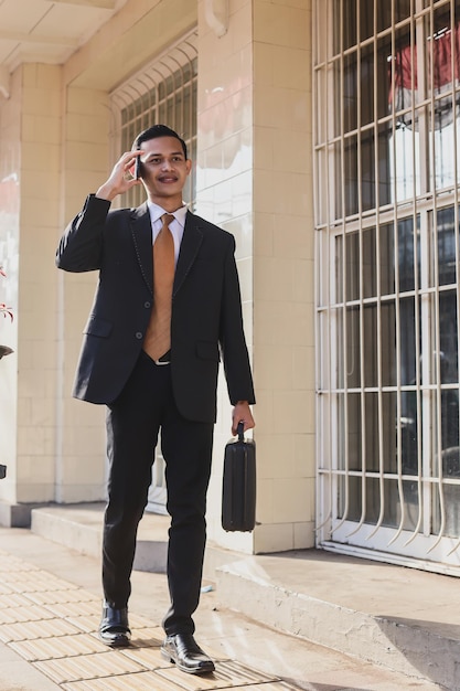 Happy young businessman with briefcase walking and talking on cell phone near business center