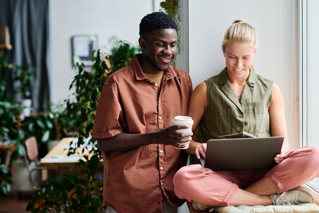 Happy young businessman with cup of coffee looking at screen of laptop