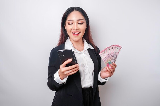 A happy young businesswoman is wearing black suit holding her phone and money in Indonesian rupiah isolated by white background