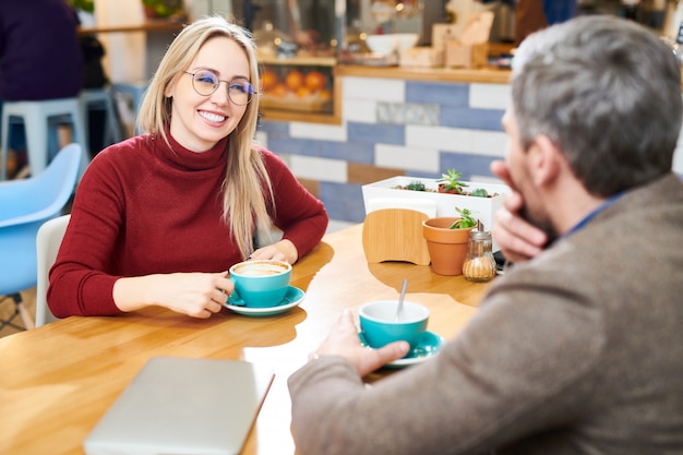 Happy young casual businesswoman having coffee by table in cafe while talking to colleague in front of her