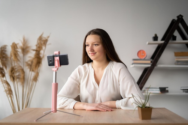 Happy young Caucasian girl takes a video on the phone while sitting at home or records her video blog smartphone on a tripod The student studies online at an Internet school blogger concept