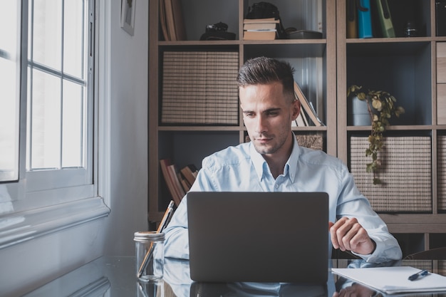 Happy young caucasian millennial businessman working at home at desk with laptop or computer having fun. Young male person in video conference taking care of the business or market.