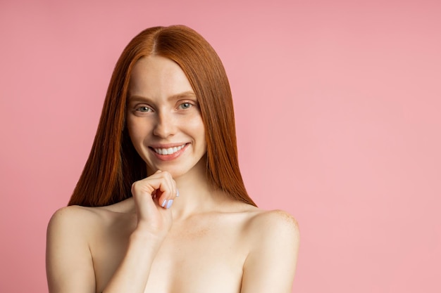 Happy young caucasian nude ginger woman with freckled clean skin touching chin, smiling pleased with result of care procedures, hair treatment posing over pink studio wall.