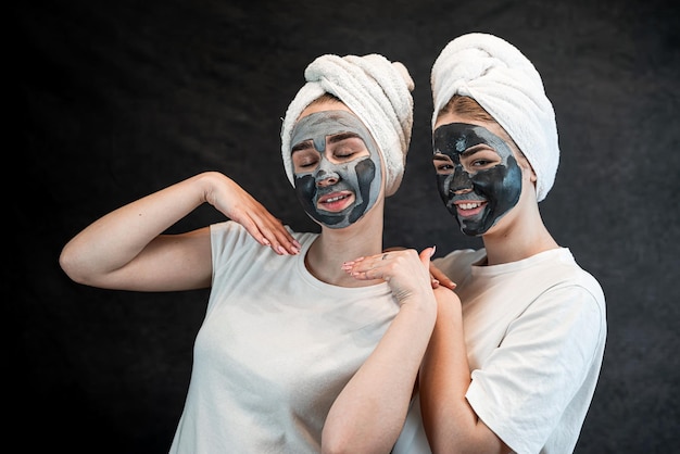 Happy young caucasian sister in hite tshirt towel wearing a charcoal mask on face isolated on black Home take care of face skin