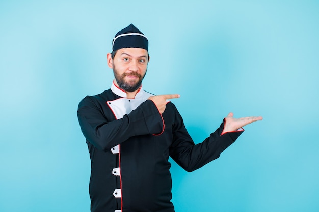 Happy young chef is pointing right side with forefinger on blue background