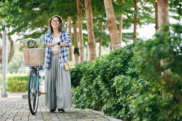 Happy young Chinese woman in plaid shirt and wide pants walking with bicycle after shopping in supermarket