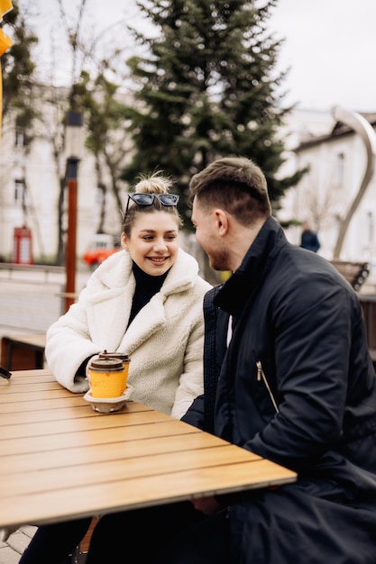 Happy young couple in love in outerwear sitting at a table outside and drinking coffee Relaxing in the open air