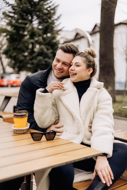 Happy young couple in love in outerwear sitting at a table outside and drinking coffee Relaxing in the open air