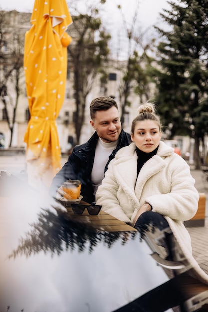 Happy young couple in love in outerwear sitting at a table outside and drinking coffee Relaxing in the open air