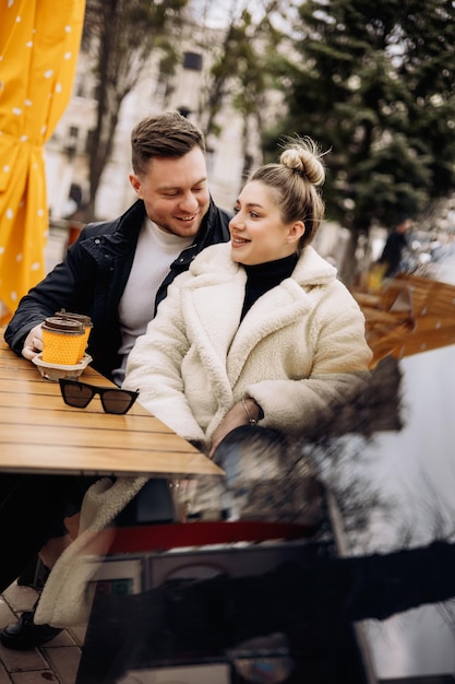 Happy young couple in love in outerwear sitting at a table outside and drinking coffee Relaxing in the open air