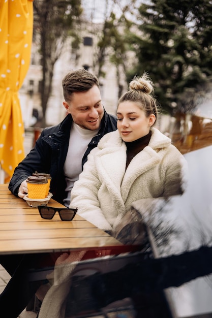 Happy young couple in love in outerwear sitting at a table outside and drinking coffee Relaxing in the open air