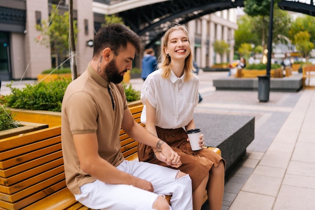 Happy young couple sitting on bench and drinking takeaway coffee enjoying time together outdoors on summer