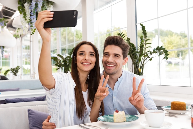 Happy young couple sitting at the cafe table, having lunch, taking a selfie