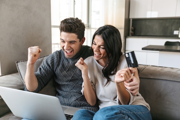 Happy young couple sitting on a couch at home, using laptop computer