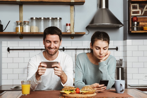 Happy young couple sitting at the kitchen during breakfast at home, using mobile phone