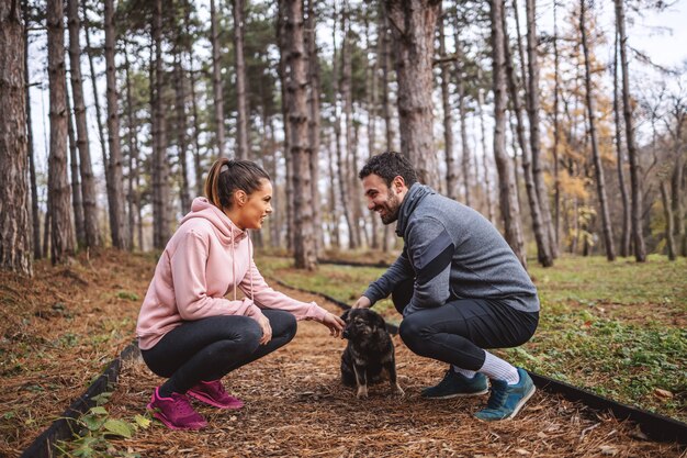 Photo happy young couple in sportswear crouching on trail in woods, looking at each other and petting stray dog. break after running.