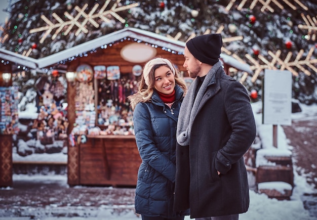 Happy young couple wearing warm clothes standing near a city Christmas tree, enjoying spending time together. Holidays, Christmas, Wintertime.